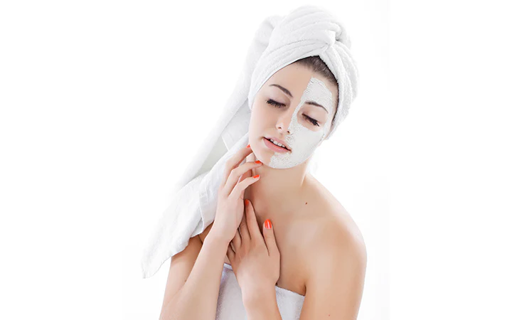 Woman with a white towel and facial mask applying skincare on her face.